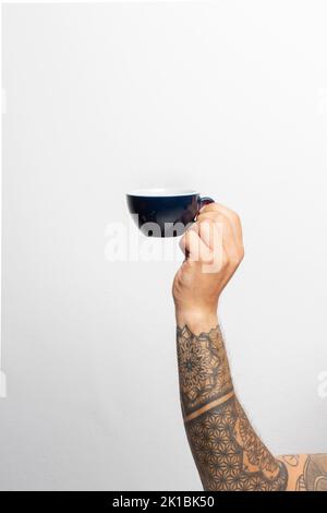 Nahaufnahme`s Arm und Hand eines tätowierten Mannes mit einer Kaffeetasse vor weißem Hintergrund Stockfoto